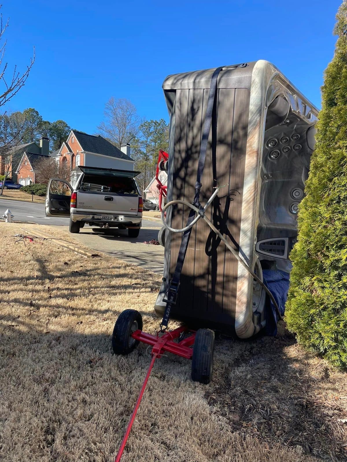 A hot tub upright on a dolly beside a hedge, moving truck in the background